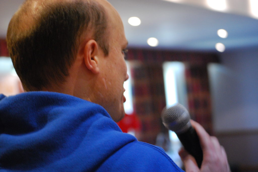 Balding man in blue hoodie speaking into a microphone