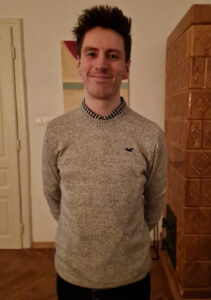 A young man with spiky brown hair and a grey jumper, standing with his hands behind his back, and smiling