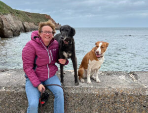 A woman with glasses, red jacket and jeans, is sitting on a coastal wall with 2 dogs, with the sea and cliffs behind her
