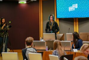 Picture of Tegan ( a young white woman with black and blue hair) standing up in the Senedd Council Chamber giving a speech