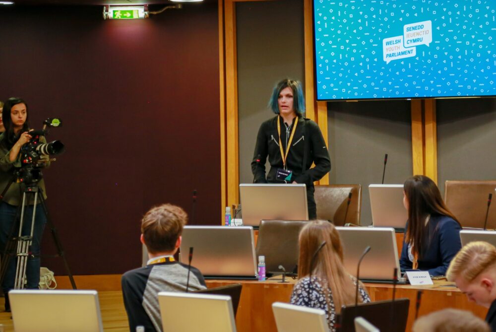 Picture of Tegan ( a young white woman with black and blue hair) standing up in the Senedd Council Chamber giving a speech