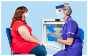 A nurse wearing a visor and holding a needle is speaking to a young woman who is wearing a mask. Behind them is a fridge full of vials