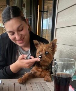 A woman with a Yorkshire Terrier sitting at a picnic table. The dog is licking some food off the woman's fingers