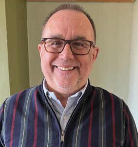 Photo of John Butterly, smiling, he is an older man with glasses, close cropped brown hair and is wearing a navy blue cardigan with red and light blue vertical stripes. 