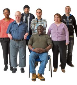 diverse group of people, all standing, one is sitting on a chair