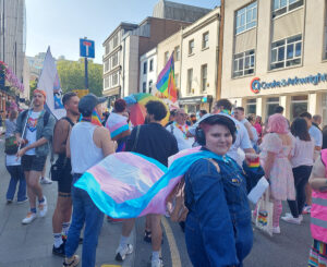 A young person wearing a blue denim boiler suit and wearing headphones, wearing the transgender flag as a cape. The flag has blue, pink and white stripes