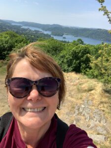 Photo of Julie outdoors,, she has bobbed red hair and is wearing sunglasses, Behind Julie is a view of a lake