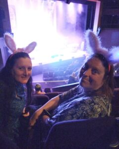 2 Gig Buddy friends at the theatre turning around and smiling at the camera, behind and below them is the stage which is bathed in white light