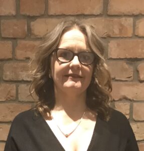 Claire has shoulder length brown wavy hair and is wearing dark framed glases.