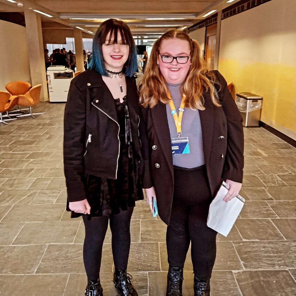 Tegan and Georgia Welsh Youth Parliament members, Tegan is dressed all in black with blue and black hair, Georgia has long hair and glasses and wearing a blazer