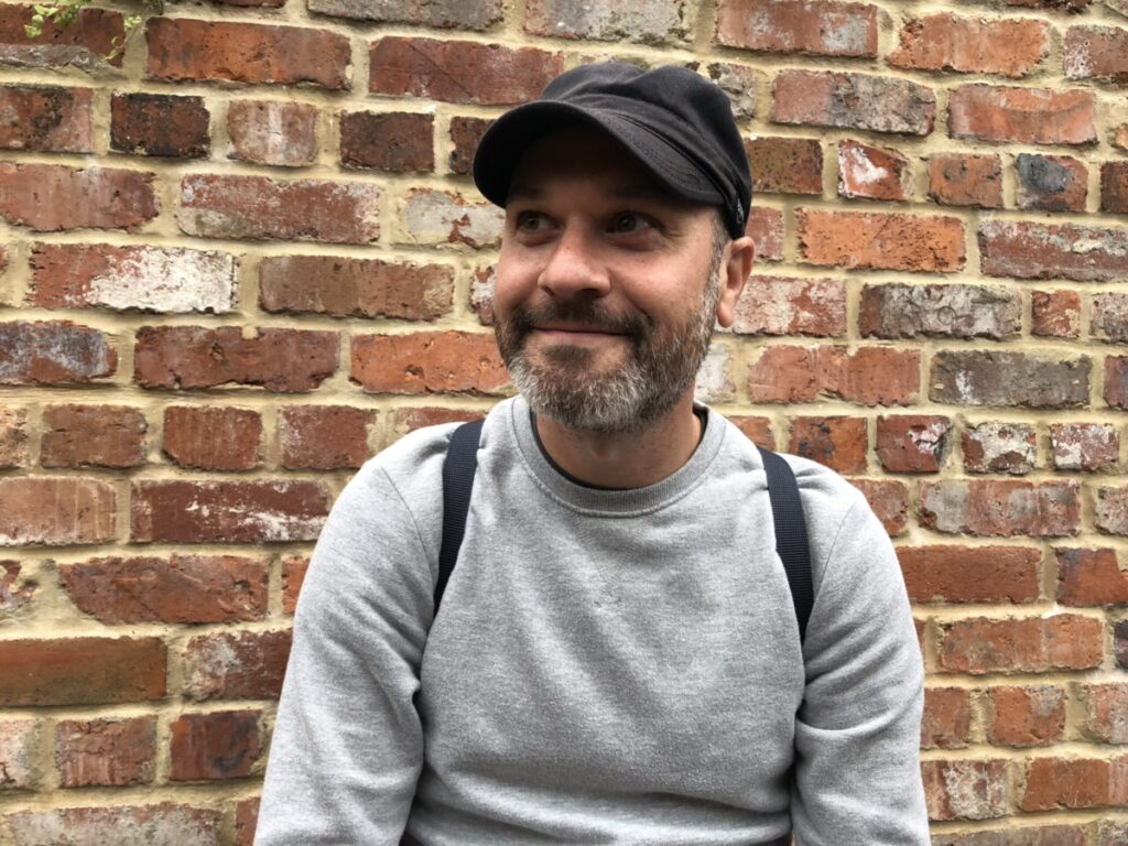 Aled has a short beard is wearing a black cap, grey jumper and is standing in front of a brick wall, looking to his right
