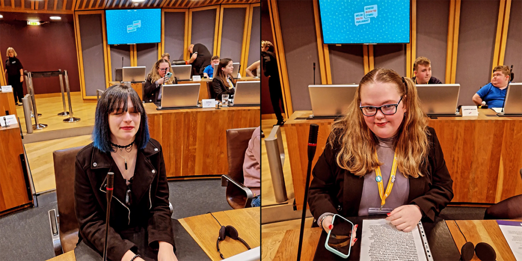 Tegan and Georgia sitting at their desks at the Welsh Youth Parliament in the Siambr debating chamber. Tegan is dressed all in black with blue and black hair, Georgia has long hair and glasses and wearing a blazer
