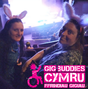 2 female Gig Buddies at the theatre wearing rabbit ear headbands and looking behind them to the camera, with the glow of the stage behind them