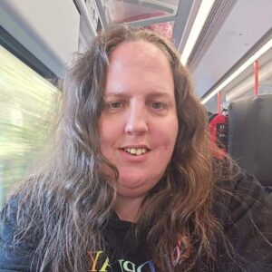 A young women with long brunette hair and a black t-shirt sitting on a train and smiling at the camera