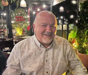 Jonathan is bald and has a short beard. He is sat is a restuarant at Christmastime.