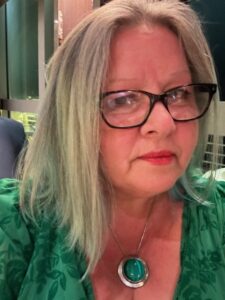 Nadine has shoulder length blond hair and is wearing dark rimmed glasses.