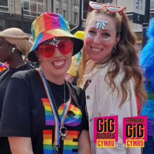 2 friends wearing Pride colours, including a rainbow hat, sunglasses and a t-shirt that says LOVE in rainbow colours, at a Pride parade