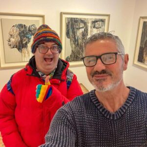 selfie of 2 friends at a gallery smiling and giving a thumbs up, framed sketches behind them