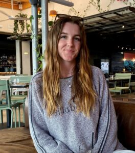 Ellie has long hair and is wearing a grey sweater and sitting in a bar somewhere sunny