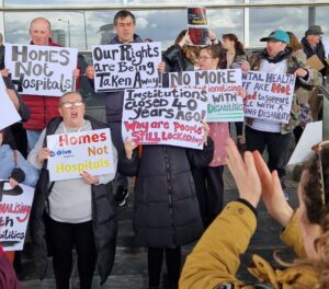 group of people with a learning disability protesting outside the Senedd and holding signs saying Homes Not Hospitals