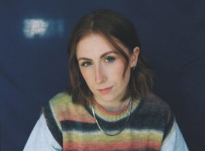 Malan has shoulder length brunette hair and is wearing a colourful and striper sweater vest