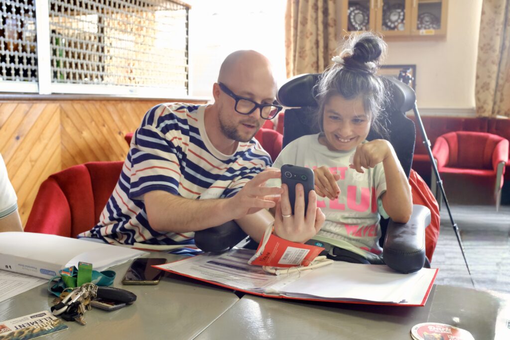 A man with a bald head, glasses and beard showing a lady with dark hair in a bun a mobile phone.