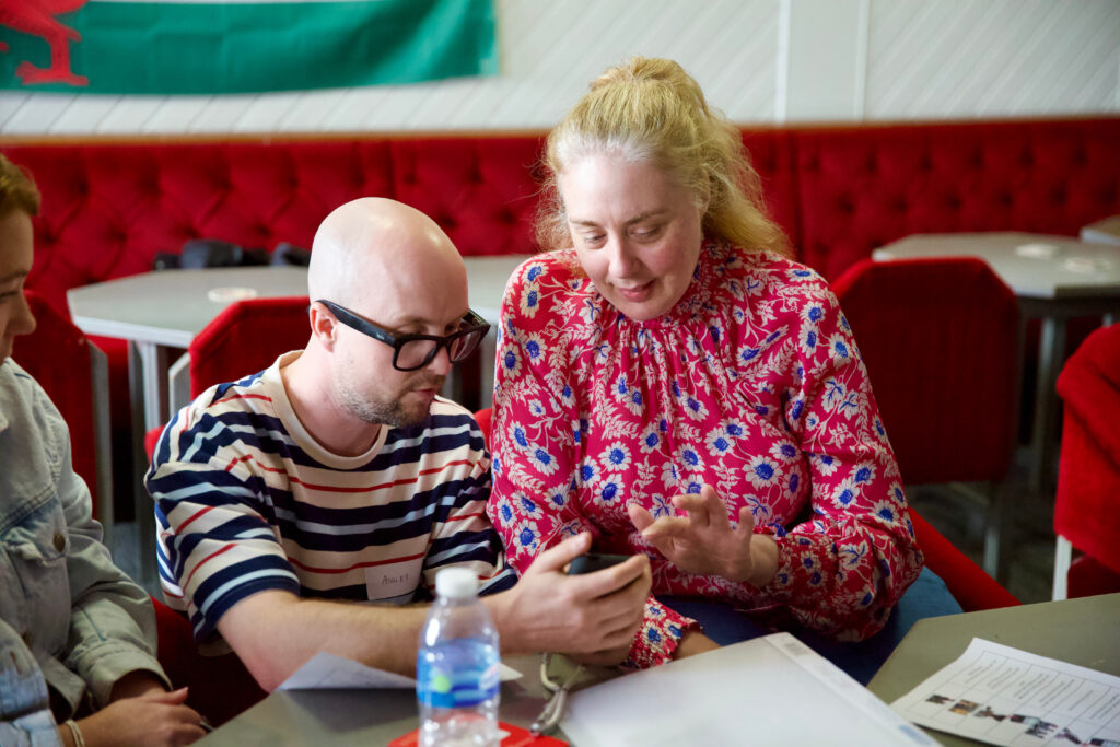 A man with a bald head, glasses and beard showing a lady with blonde hair in a ponytail how to use a mobile phone.