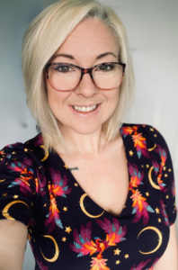 A person with short blonde hair and glasses smiling at the camera. Wearing a patterned top.