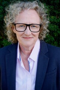 A smiling person with blonde curly hair, wearing glasses, a pink shirt and blazer.
