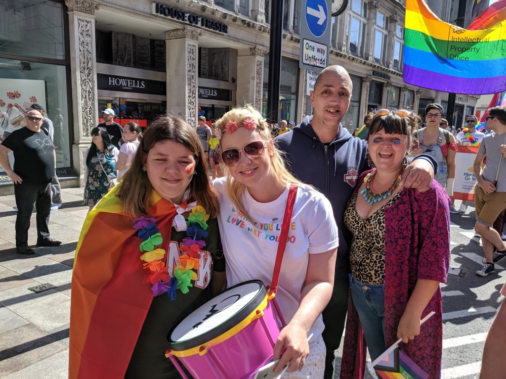 A group of Gig Buddies during the Pride Cymru parade. They are smiling and are wearing lgbt rainbows and have an lgbt rainbow flag
