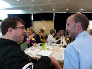 Two young men in a meeting facing each other and smiling, behind them other people are having discussions