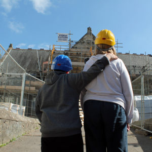 Two people with their backs to the camera are looking at a building site. One person has their arm around the shoulder of the other.