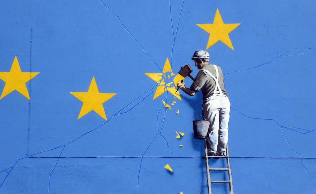 Man in overalls on a ladder removing one of the yellow stars from a gigantic EU flag. Mural is by Banksy