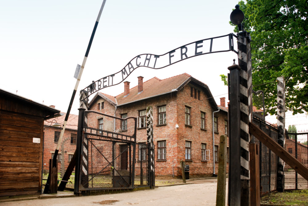 entrance to Auschwitz 1 - barrier is raised next to an iron sign over the entrance that reads in English Work Sets You Free. Behind the barrier are rows of large red bricked buildings