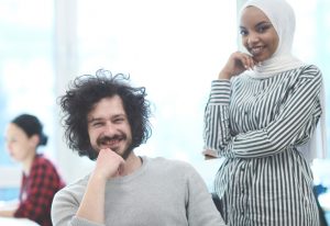 2 young people in a office, a young man with a beard and grey jumper is sitting and smiling, next to him is a young woman in a stripy black and white dress and hijab 