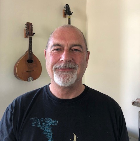 A close up of Adrian, a light skinned man with a grey goatee beard, wearing a black tshirt. Behind him on the wall is hanging a wooden mandolin.