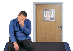 Man looking distressed, sat on crash mat in locked room with staff looking through a small window in the door.